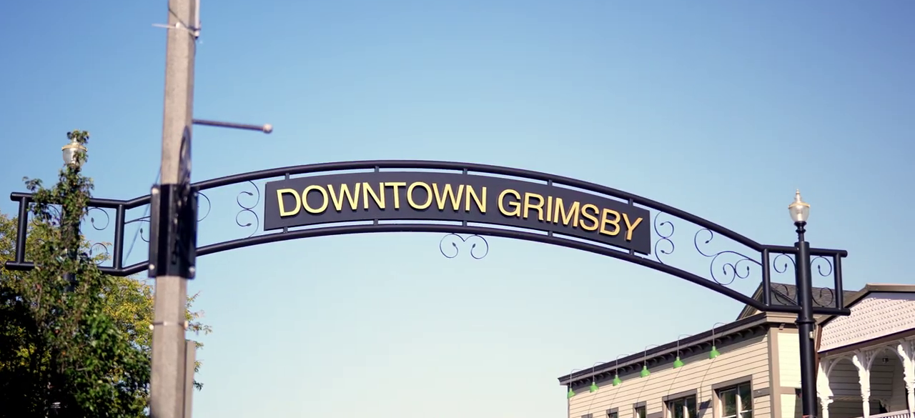 View over Grimsby from the Niagara Escarpment, with the Downtown Grimsby arch sign, vineyard rows on the slope, rooftops below and Lake Ontario on the horizon, and a set of house keys on a rock in the foreground