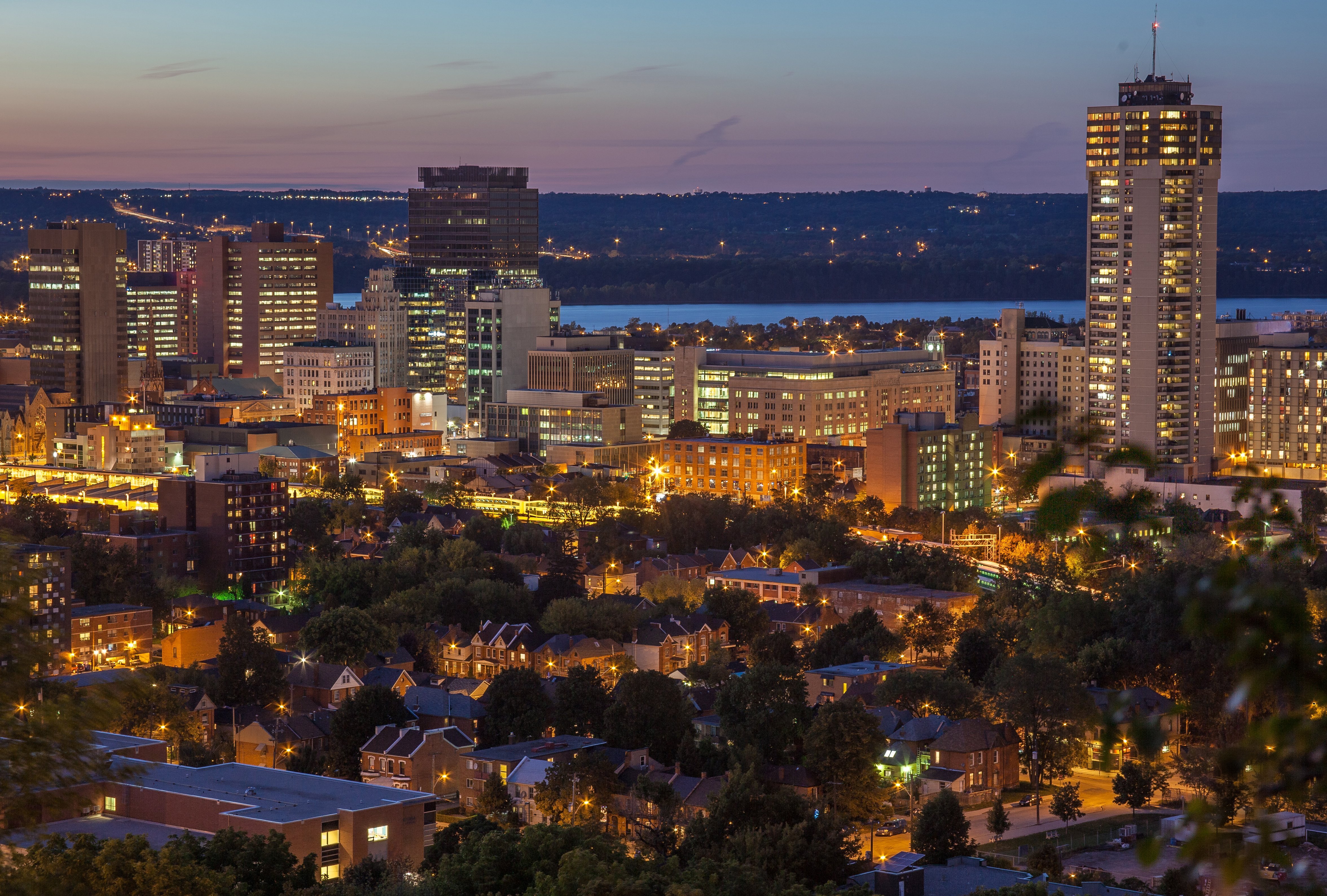 Glass and dark-metal high-rise condo tower lit up at dusk in downtown Hamilton, seen from street level with the city skyline behind it